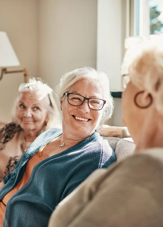 three senior women sit on a couch at home and smile during conversation