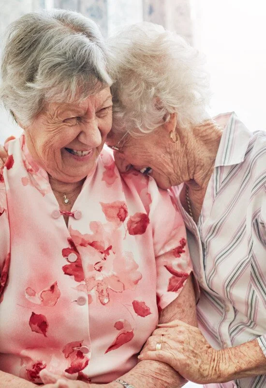 two senior women giggle, with one leaning to rest her face on the other's shoulder