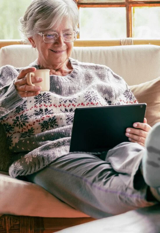 senior woman relaxes on couch with her feet up, reading on a kindle and drinking tea