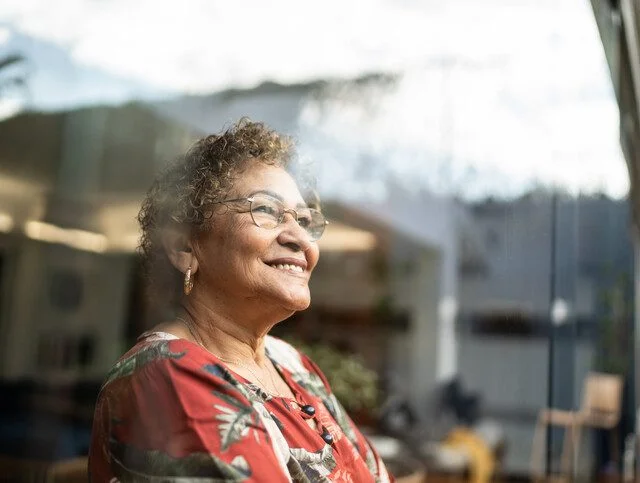 senior woman in glasses and patterned blouse smiles while looking out of a window