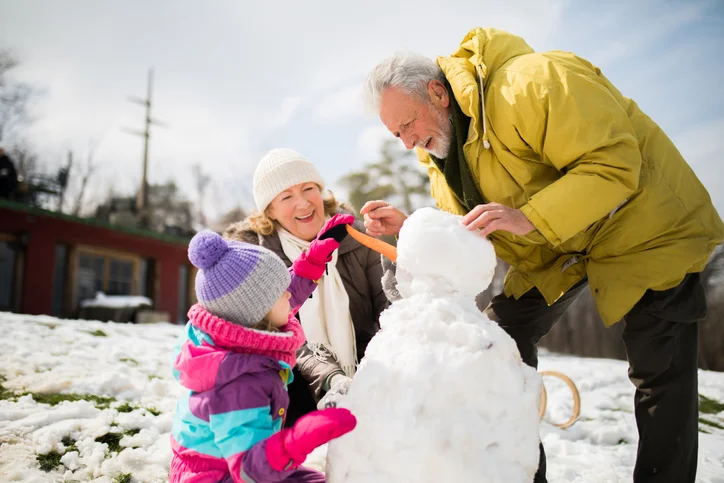 senior couple in winter coats smile while helping their granddaughter build a snowman