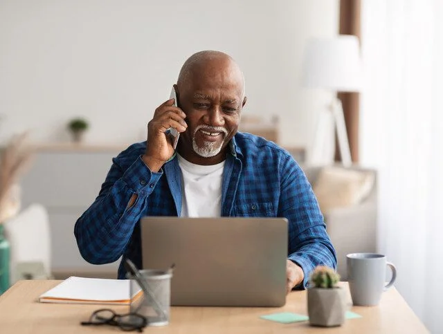 senior man sitting at his table on the phone, looking at his laptop and smiling