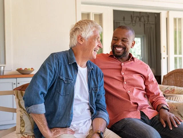 two senior men smile and sit on a couch together