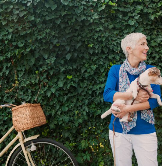 senior woman holds her dog while standing next to her vintage bike, backdropped by wall of ivy